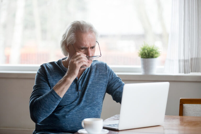 Shocked frustrated senior man taking off glasses looking at laptop